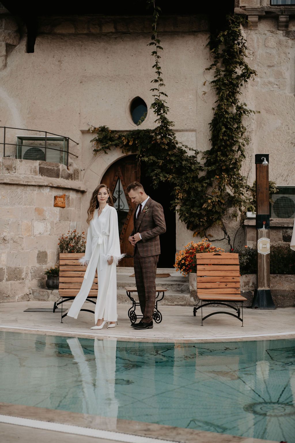 Elegant couple in wedding attire standing by a pool at a rustic stone venue with ivy-covered walls in Cappadocia.