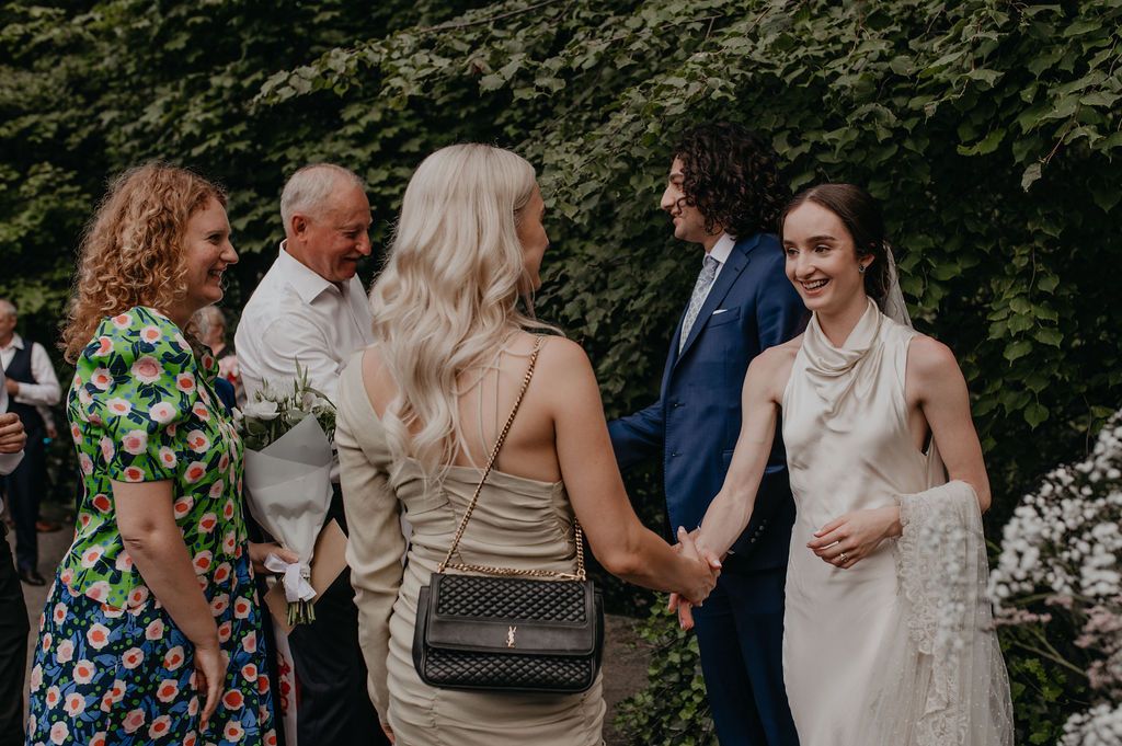 Bride in white dress and pearls smiles while holding hands with guests at an outdoor wedding reception.
