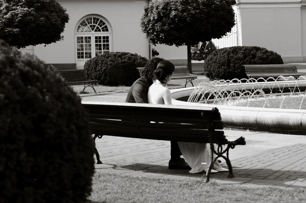 Couple sitting close together on a park bench near a fountain, surrounded by trimmed hedges, in black and white.