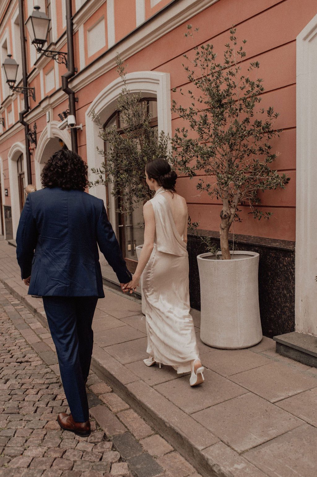 Bride and groom walking hand in hand along a cobblestone street past a pink building on their wedding day.