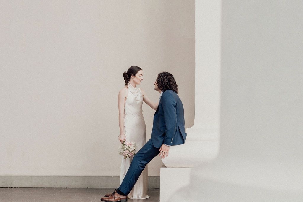 Bride in beige dress holding flowers faces groom in navy suit seated on white bench against minimalist wall.