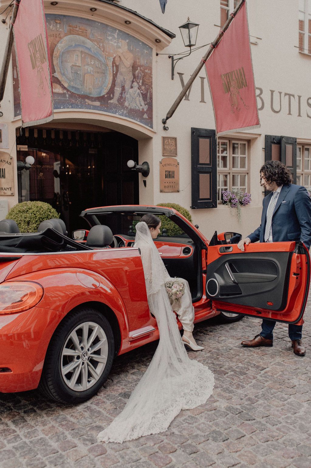 Bride in lace veil exits red convertible as groom holds door outside European venue.