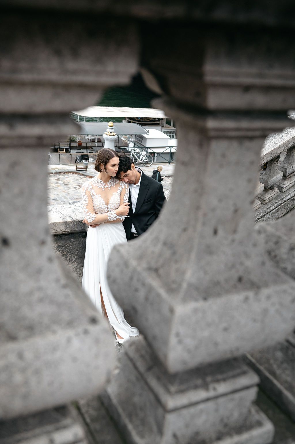 Bride in lace gown and groom in black suit share intimate moment framed by stone architecture at outdoor wedding venue.