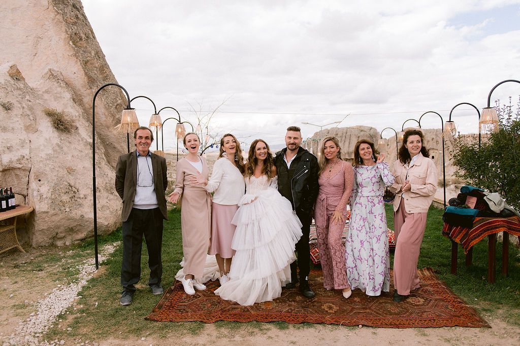 Wedding party posing outdoors near cave rock formations in Cappadocia, bride in white gown with guests in pink and floral attire.