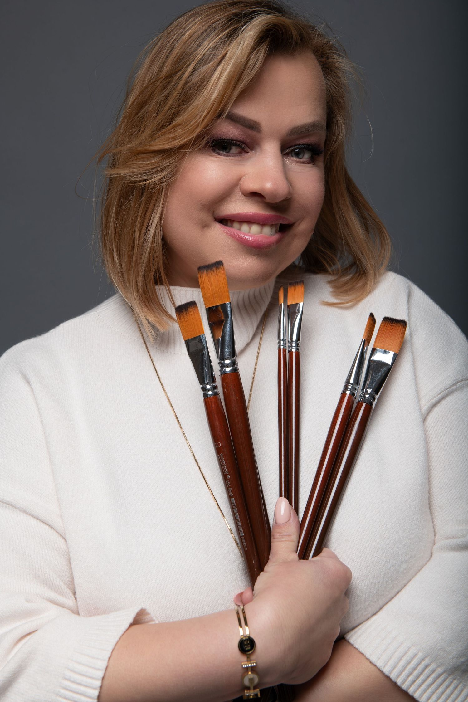 Smiling woman in white sweater holding multiple artist paintbrushes, posing against a gray background.