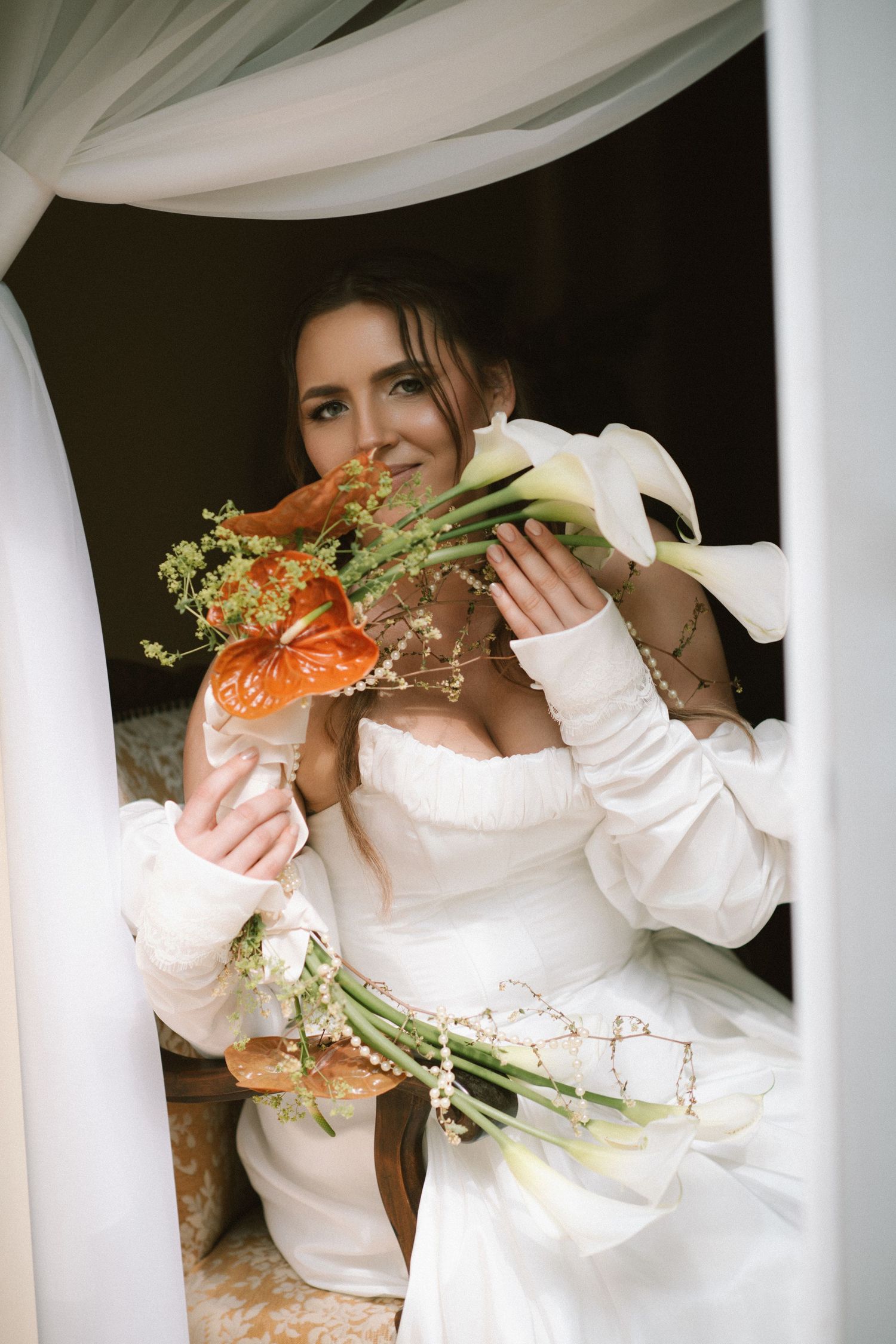 Bride in white off-shoulder gown holds colorful wedding bouquets with calla lilies and orange anthuriums, smiling softly.