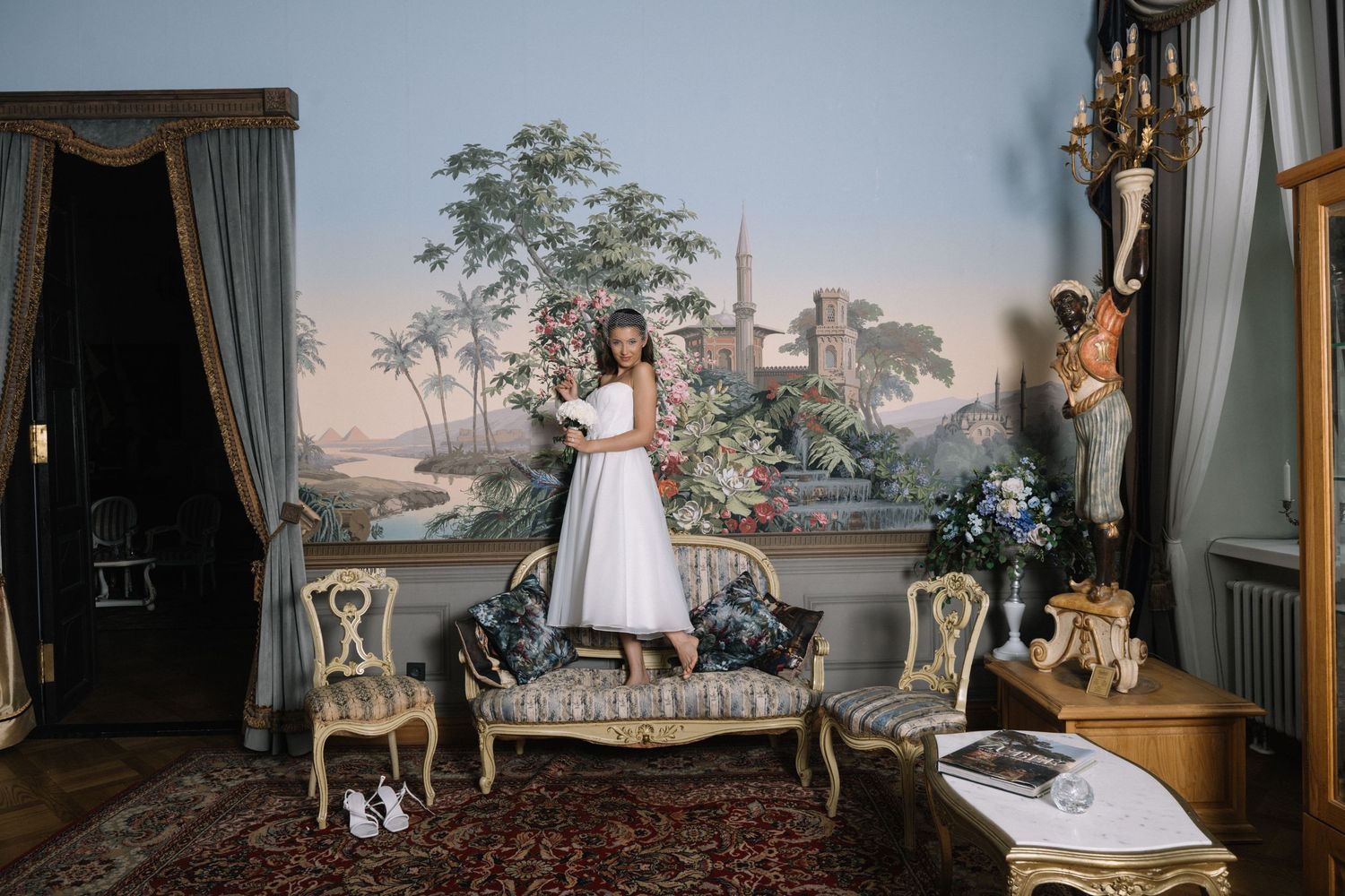 Woman in white dress standing in an elegant room with ornate furniture and scenic wall murals.