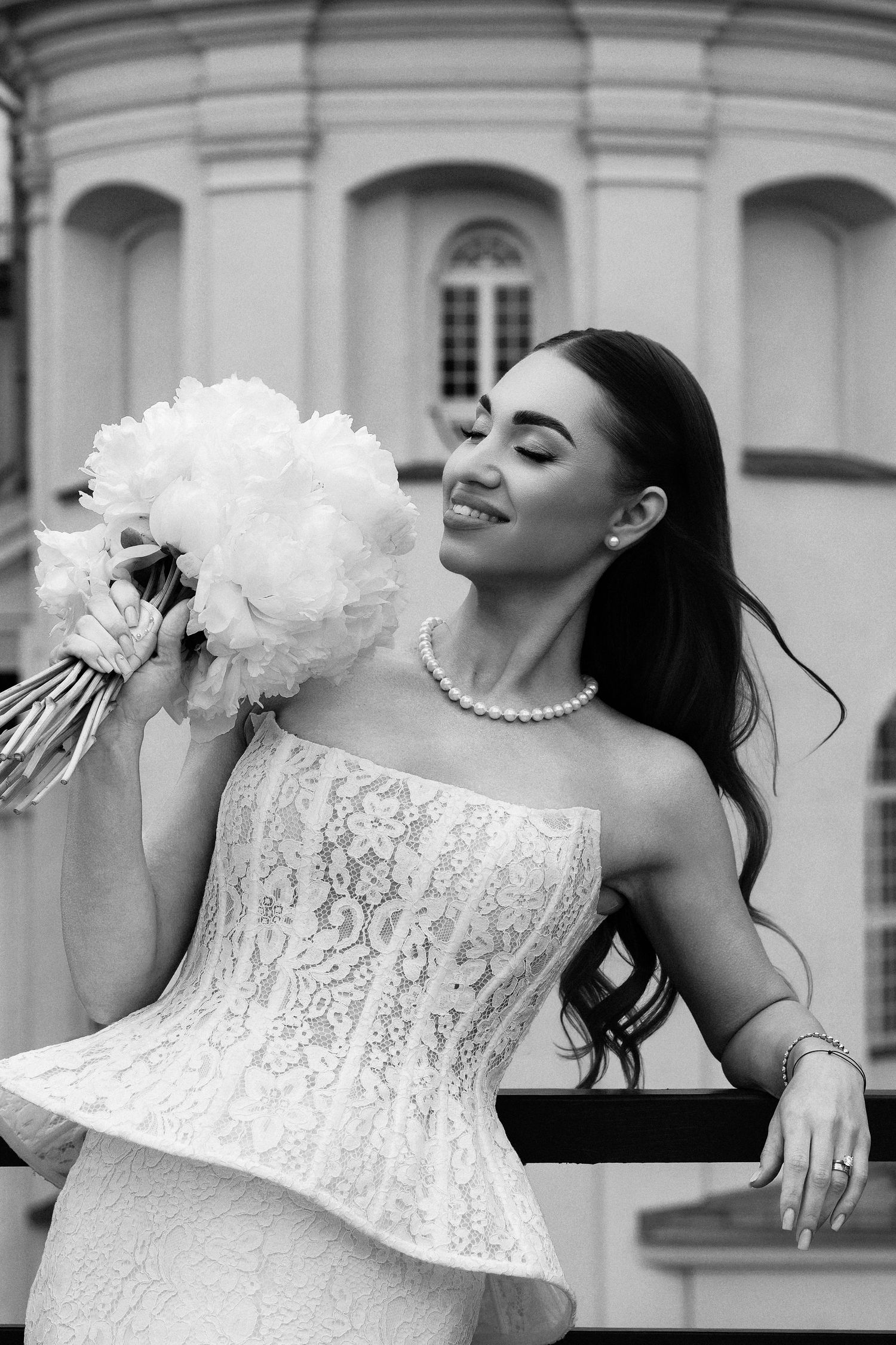 Black and white photo of a smiling bride in lace gown holding a peony bouquet, wearing pearl necklace outdoors.