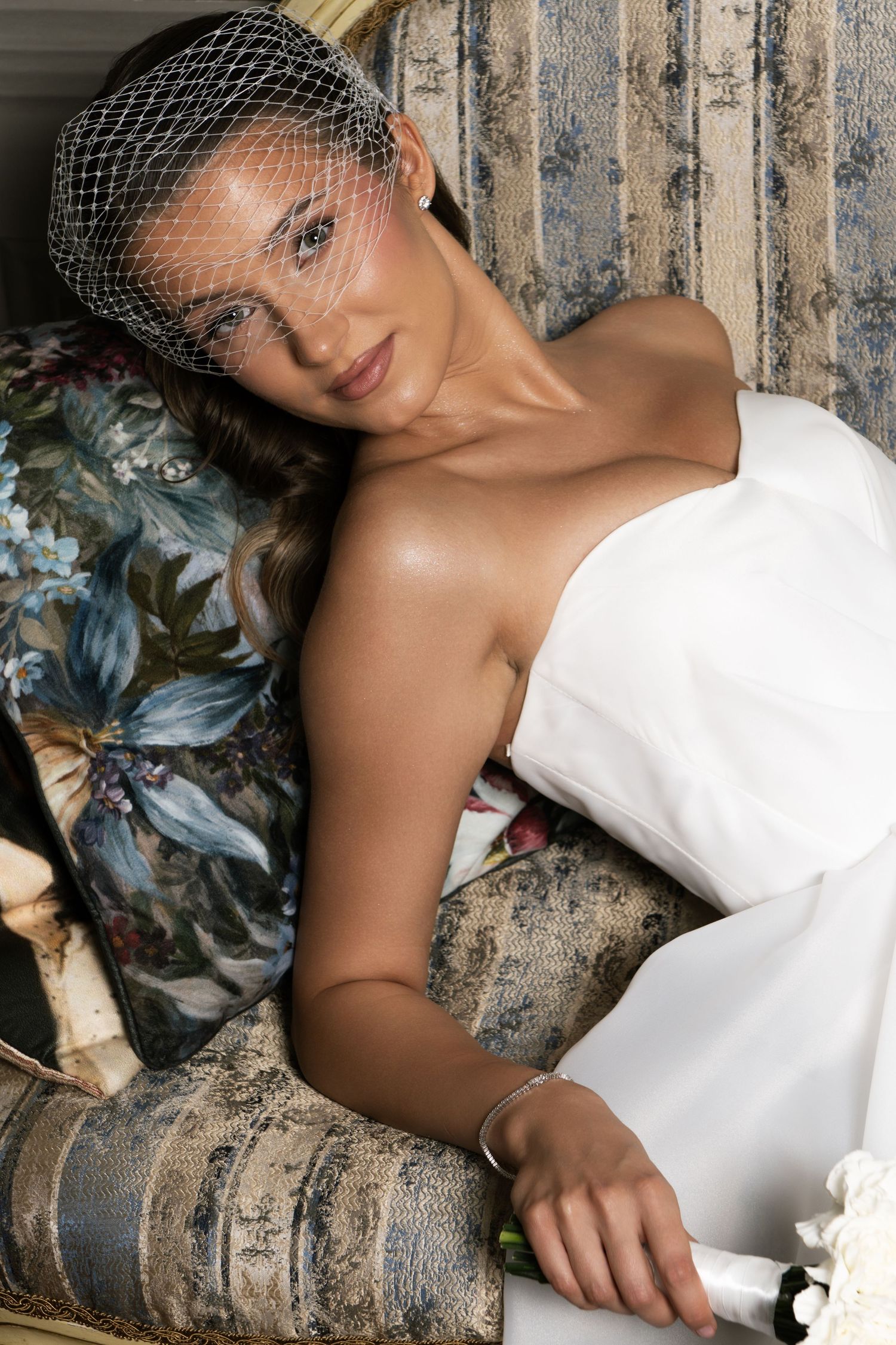 Elegant bride in white strapless gown and birdcage veil reclining gracefully on a floral upholstered chair.