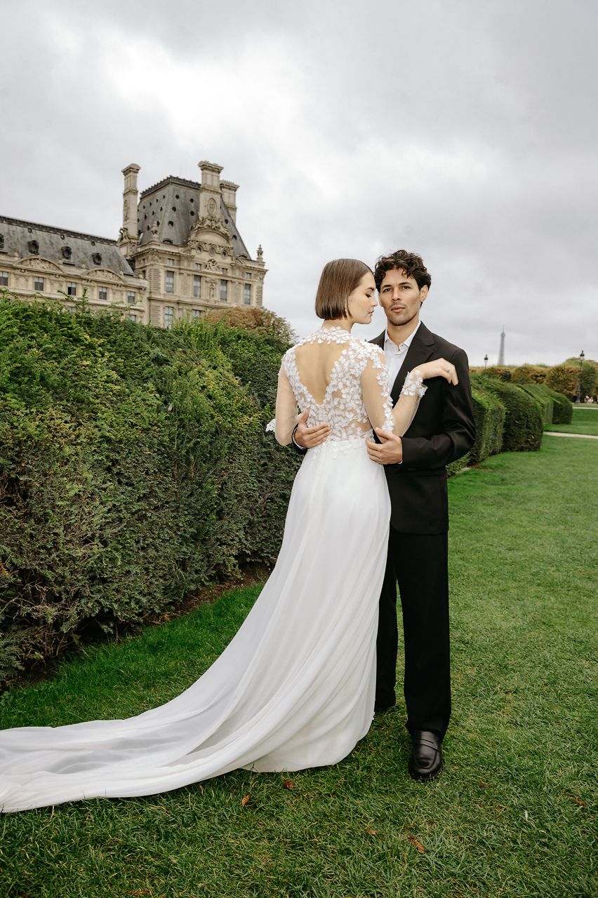 A couple in wedding attire embraces on a lush green lawn with a historic stone building in the background.