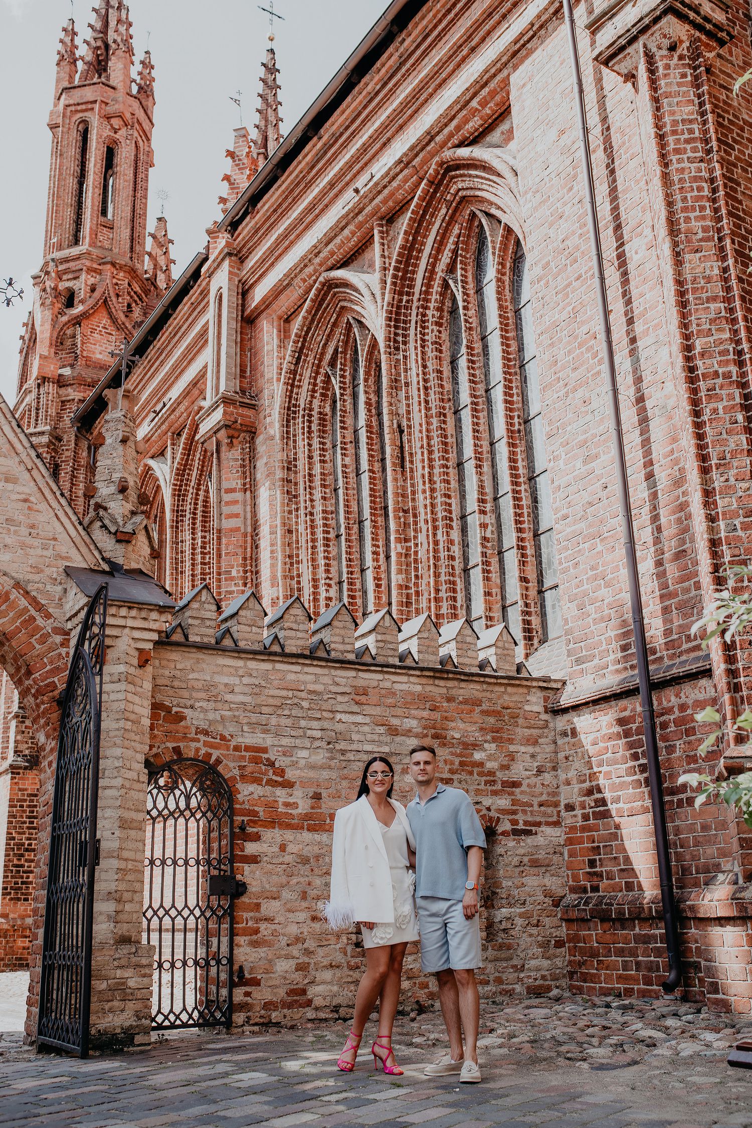Couple stands in front of ornate Gothic church architecture with red brick walls and spired tower in Vilnius, Lithuania.