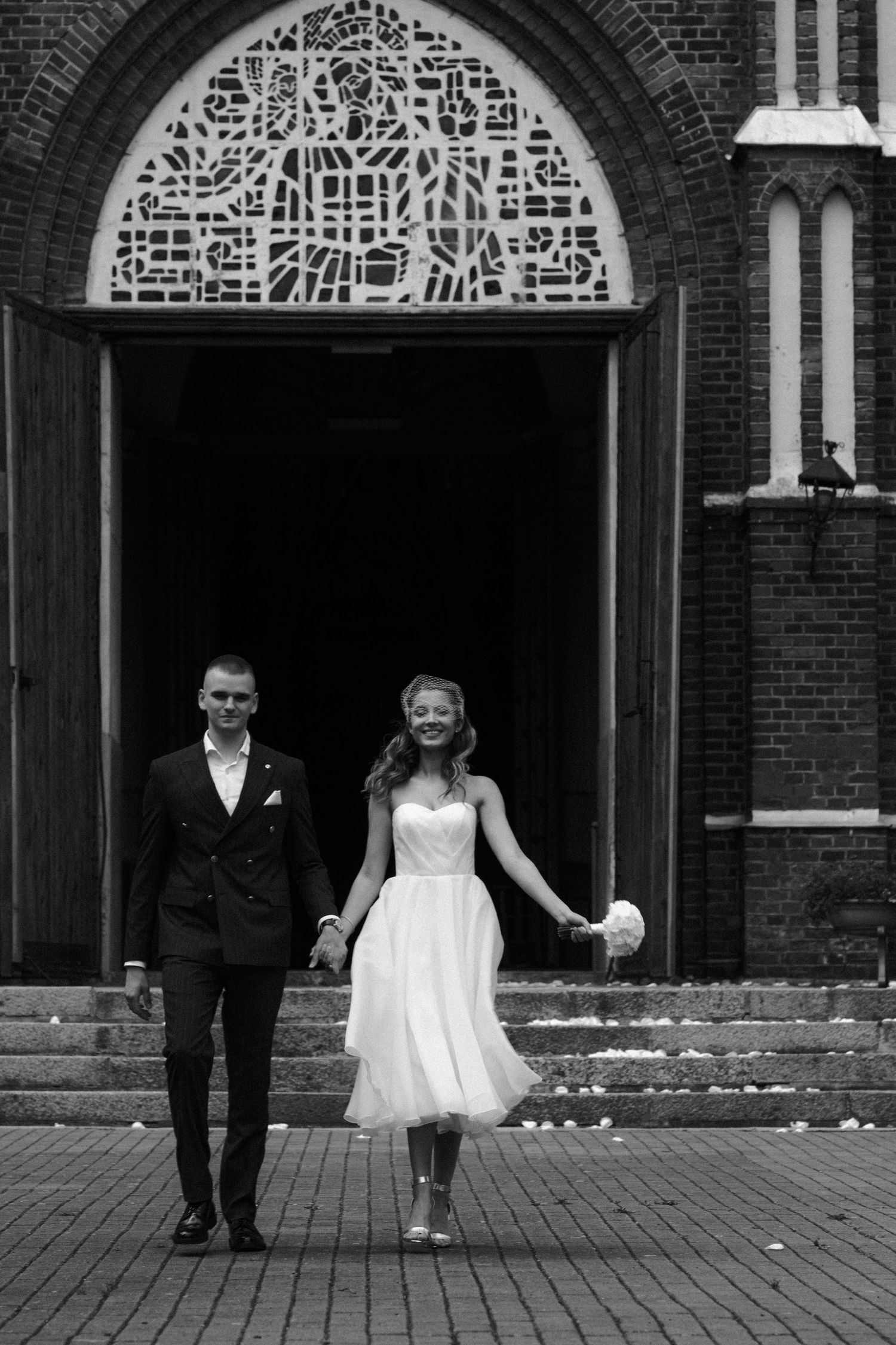 Black and white photo of a smiling bride and groom holding hands outside a brick church after their wedding ceremony.