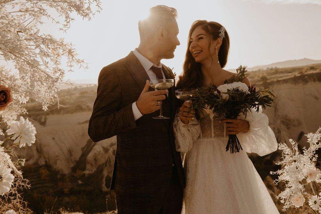 A smiling bride and groom toast with glasses outdoors at golden hour, she holds a bouquet in a white gown.