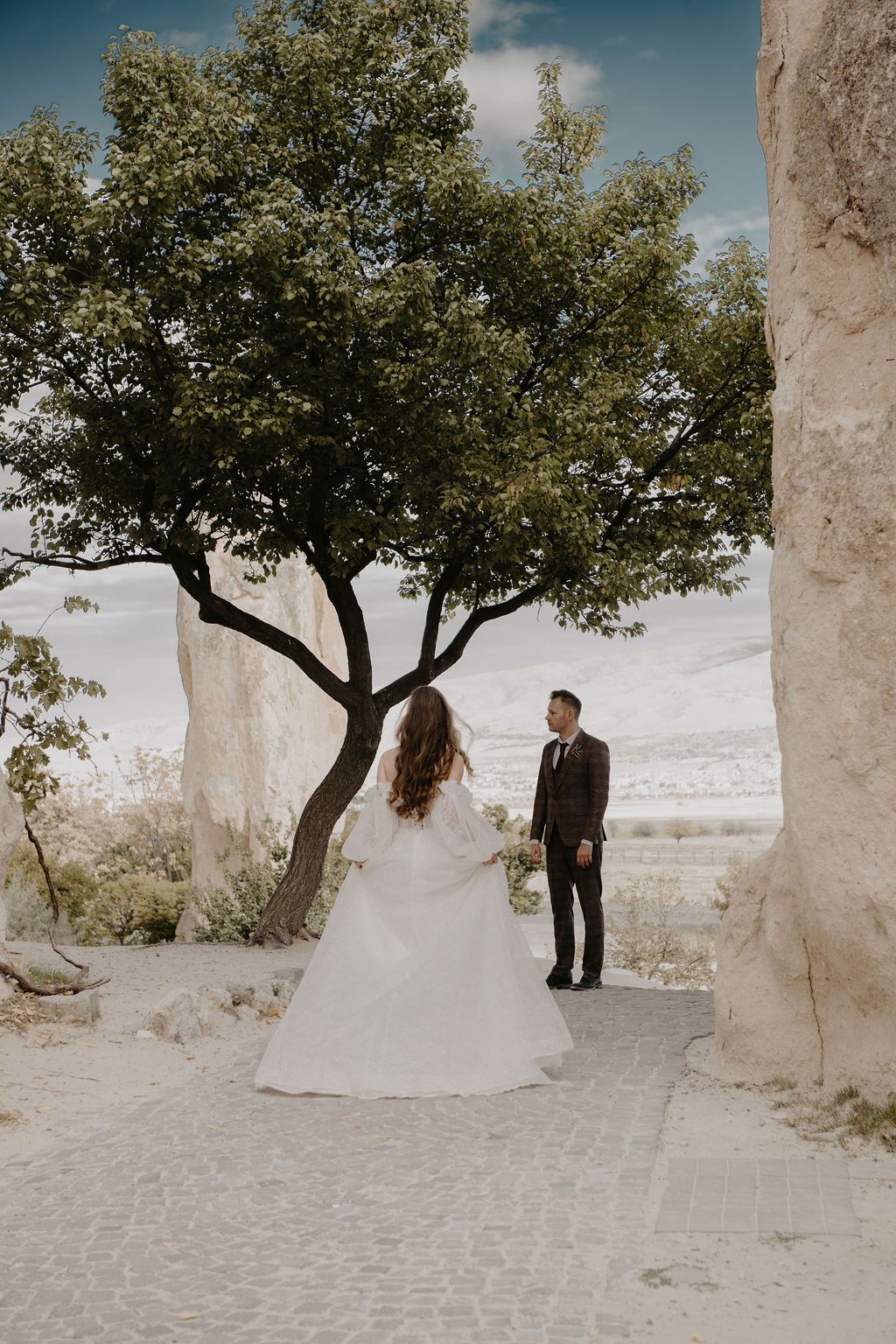 Bride and groom share a romantic moment under a tree amid rocky landscape during an outdoor wedding ceremony.