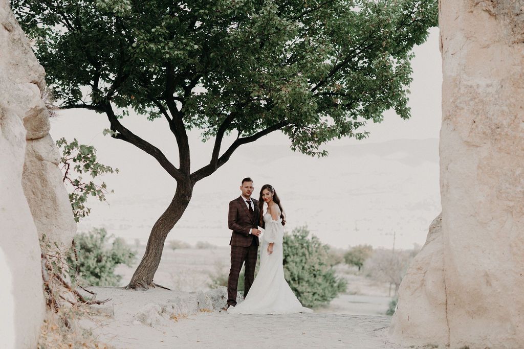 Bride and groom pose under a tree amid rocky landscape on their wedding day.