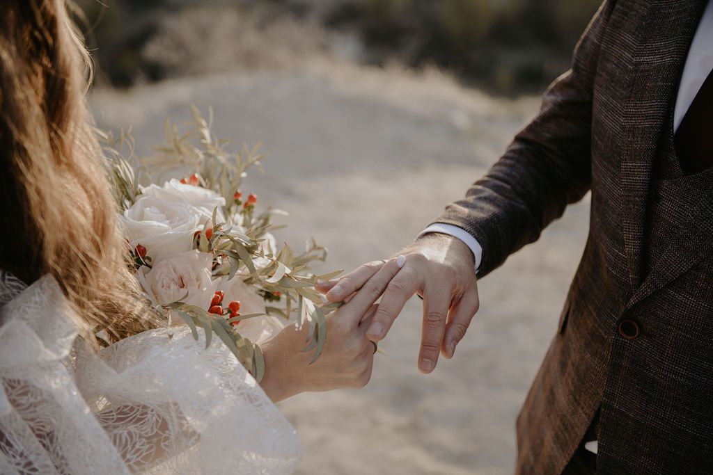 Bride and groom exchanging rings outdoors, bride holding a boho floral bouquet with white and red blooms.