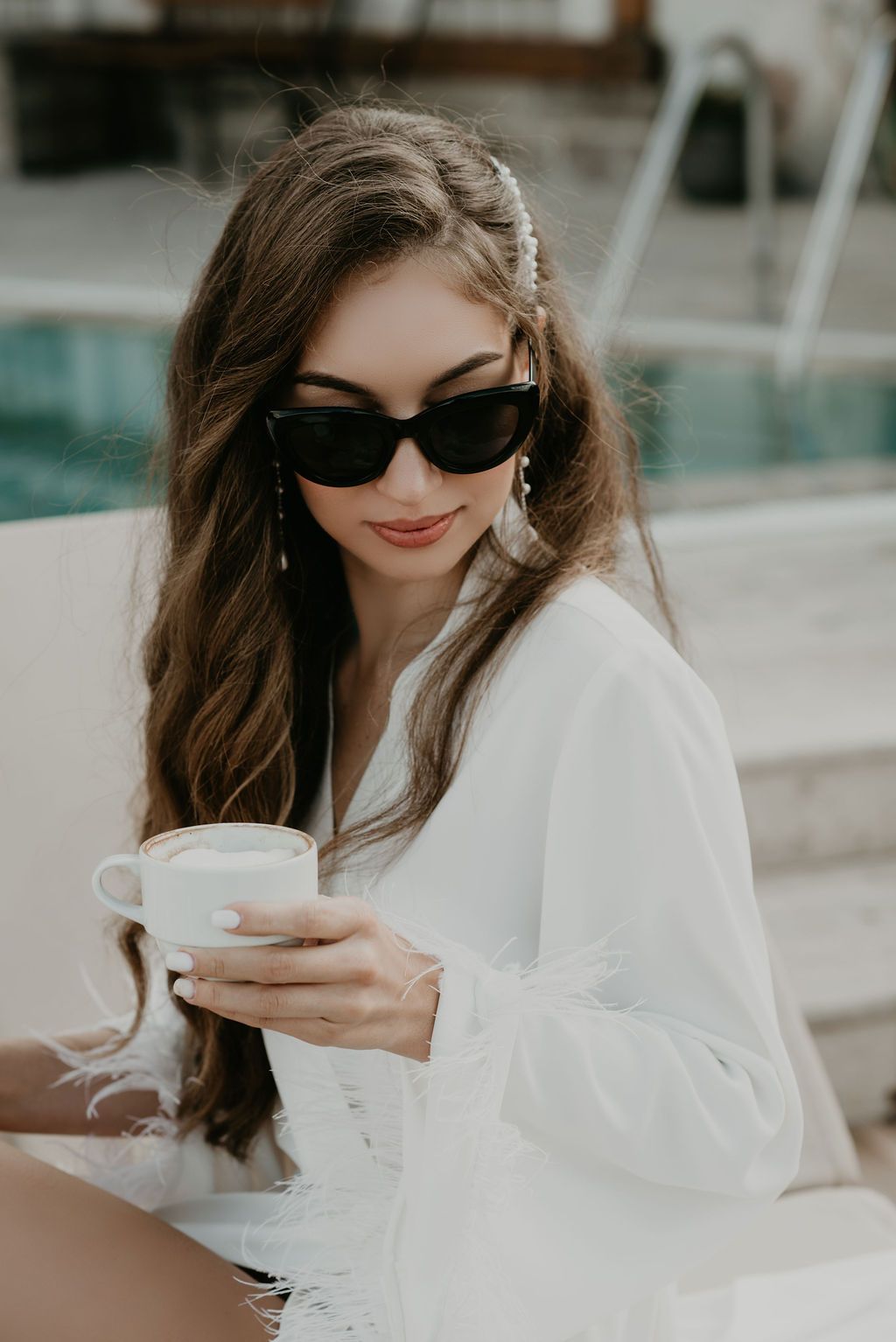 Woman in white blouse and black sunglasses holding a latte cup outdoors near a pool.
