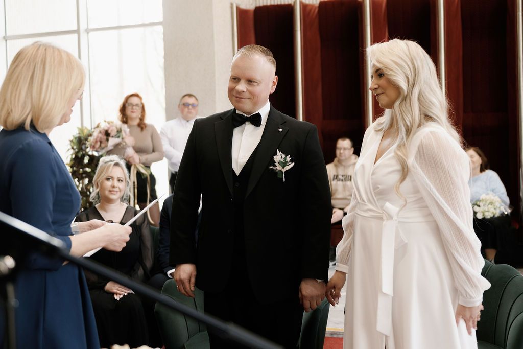 Bride and groom exchanging vows at elegant wedding ceremony, officiant reading from script, guests seated in background.