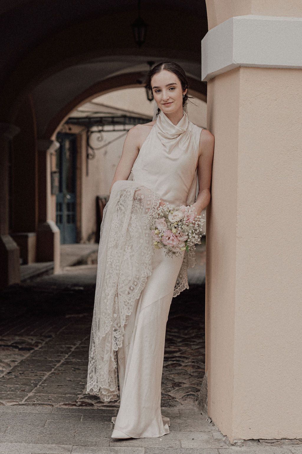 Bride in elegant ivory satin halter gown with lace wrap holding pink bouquet, leaning against a column.