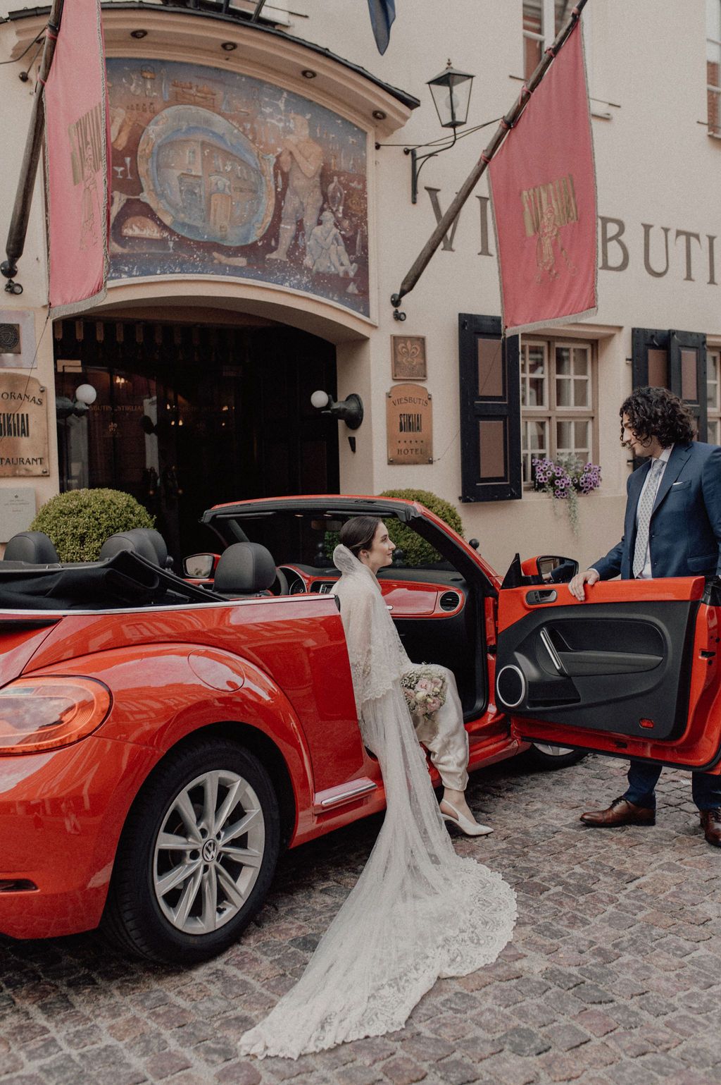 Bride in lace gown exits red VW Beetle convertible as groom watches outside a European venue.