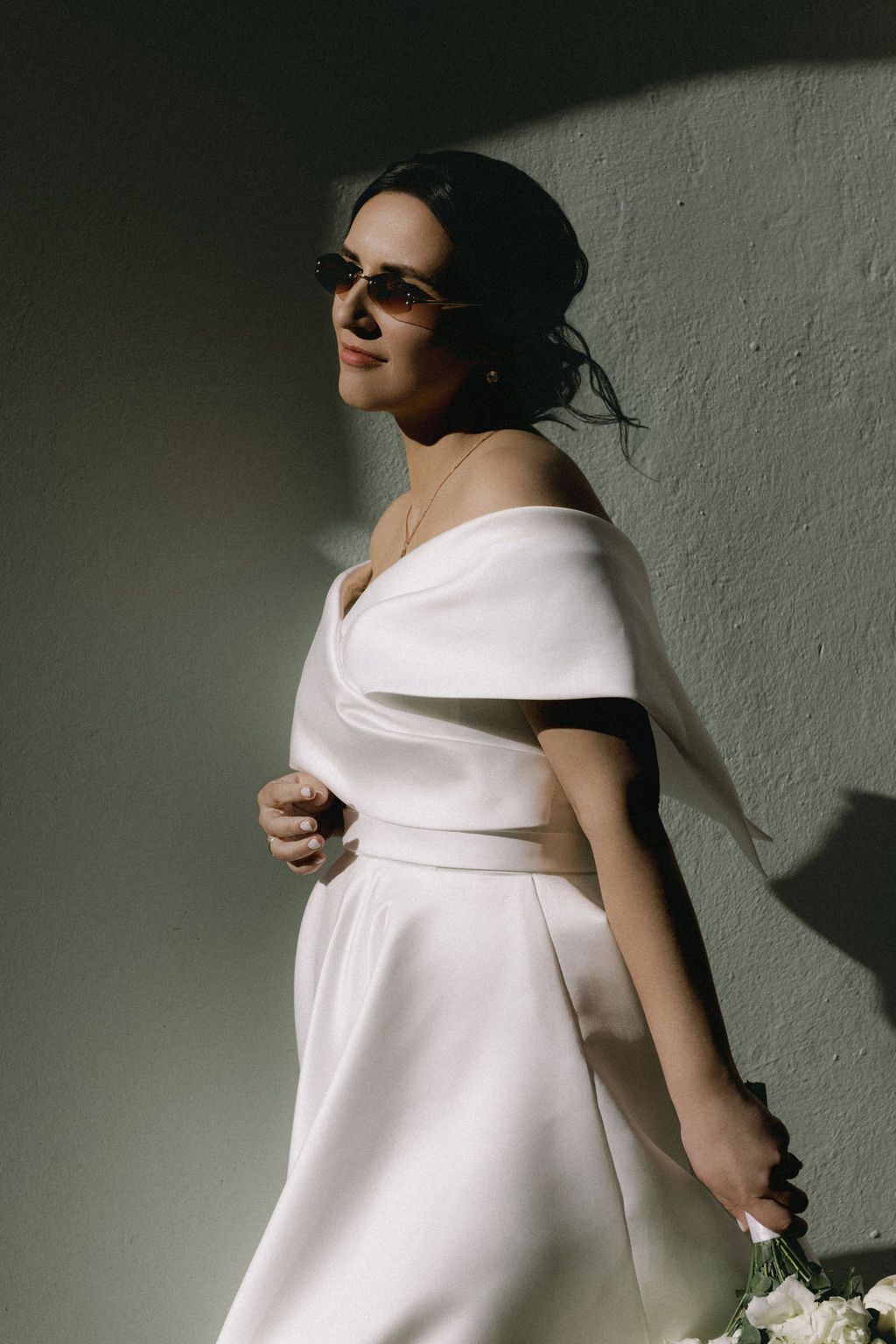 Elegant bride in white off-shoulder gown and sunglasses poses against a textured wall holding a white bouquet.
