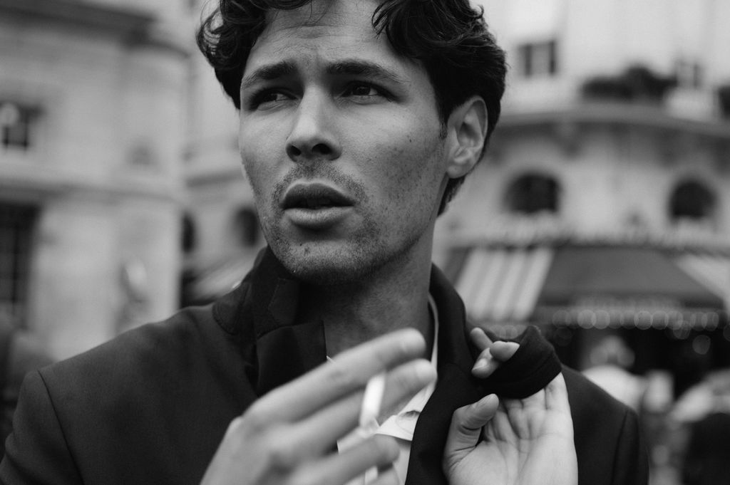 Black and white portrait of a stylish man holding a cigarette on a bustling city street.