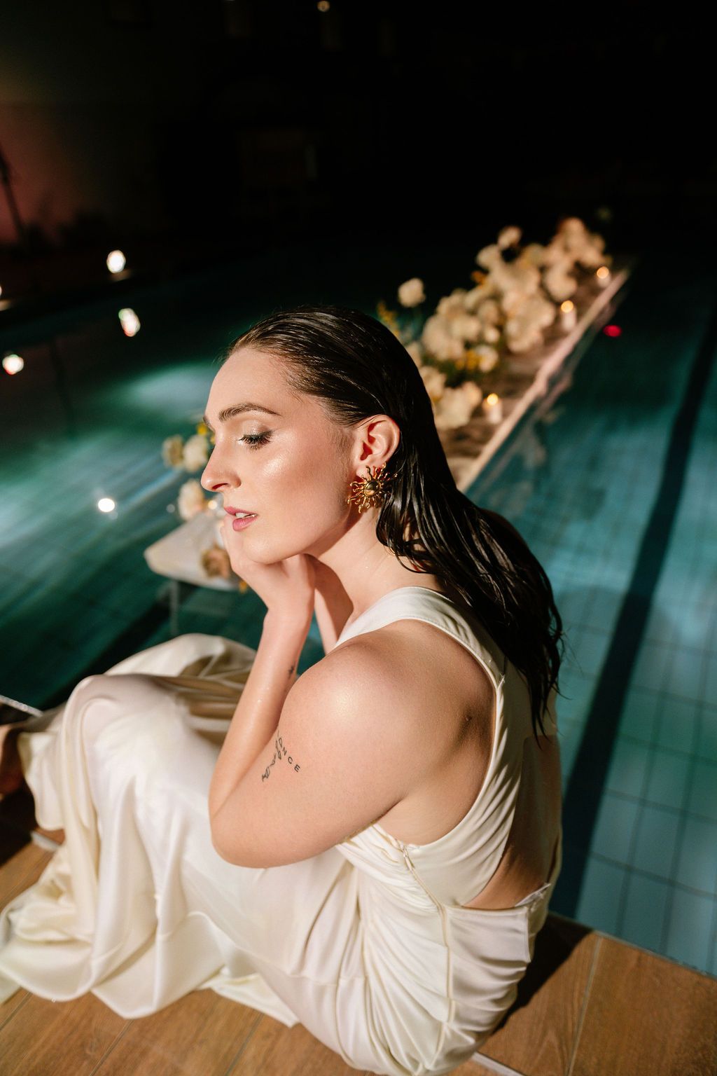 Elegant woman in white satin dress with gold earrings sitting poolside near floral arrangement at night.