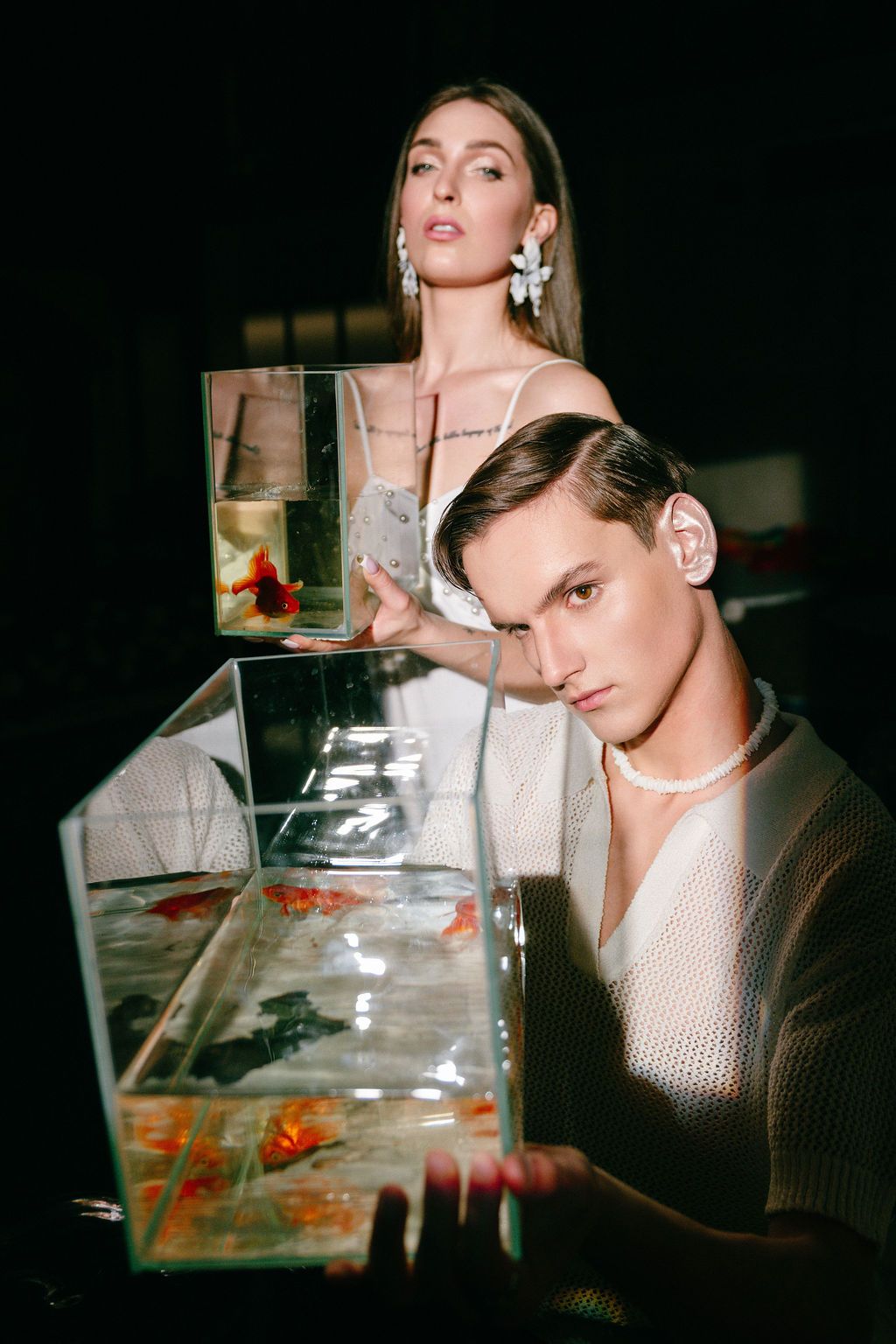 Two models pose with goldfish tanks in a dark studio, woman wearing statement earrings, man in knit top with pearl necklace.