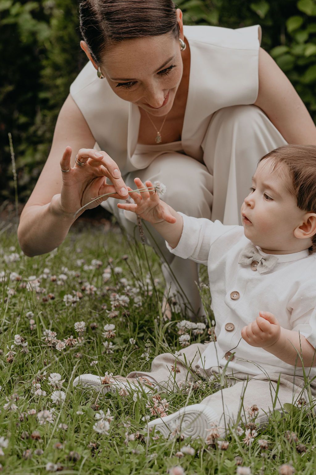 A bride in white crouches in a flower meadow, joyfully interacting with a baby dressed in white.