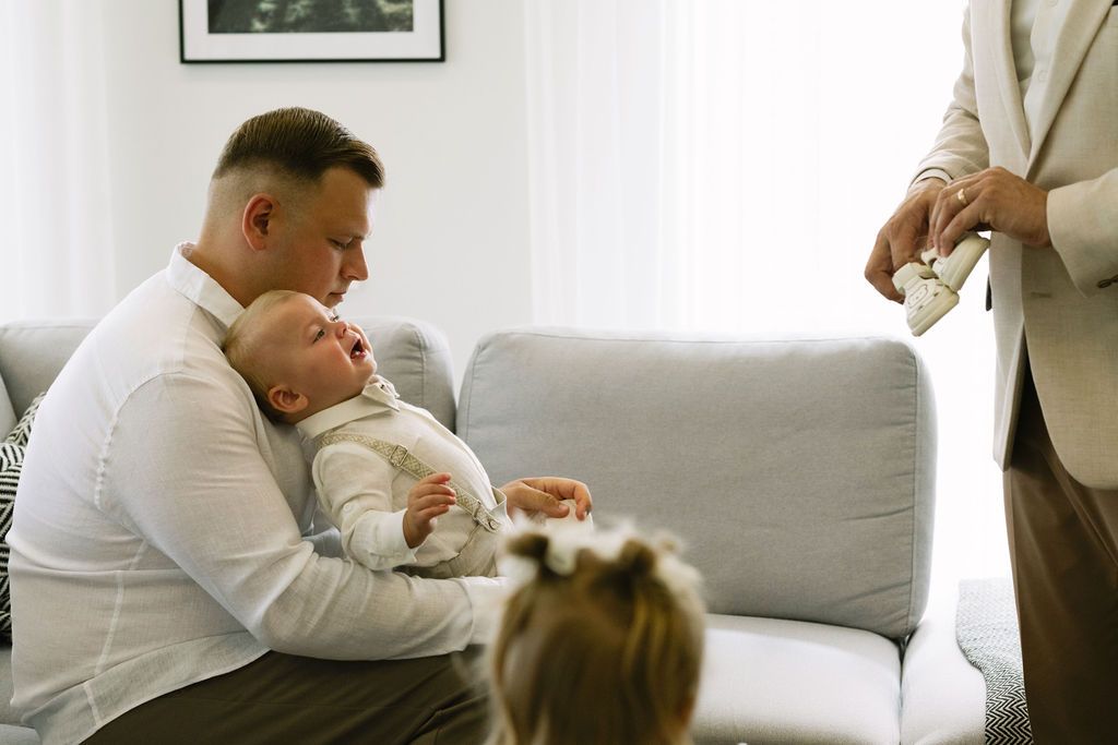 A father cradles his newborn baby on a gray sofa while family members gather around in a bright white room.
