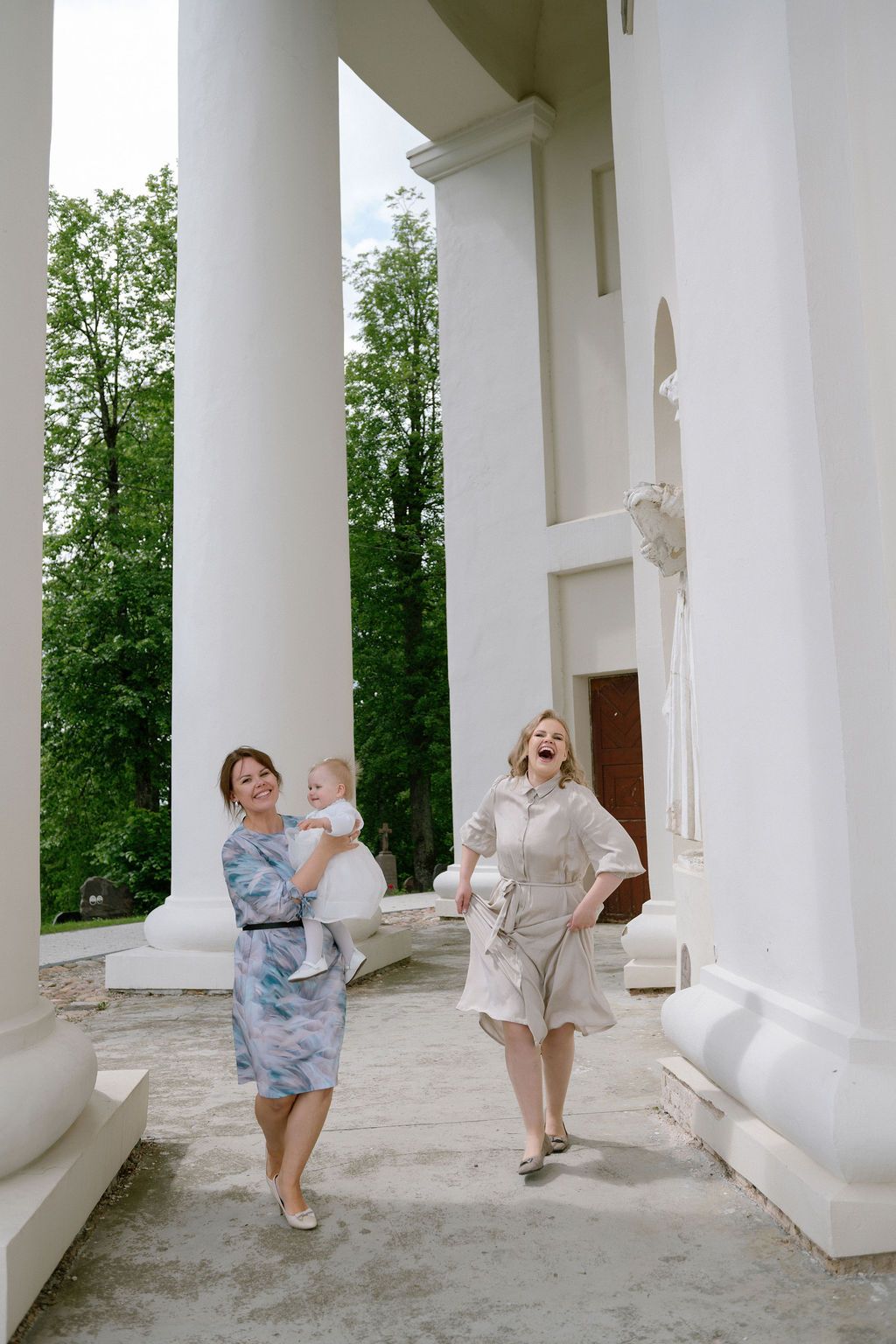 Two women with baby walking among tall white columns outside a neoclassical building.