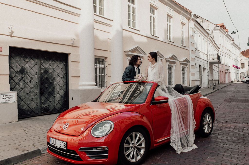 Couple in wedding attire sitting on a red VW Beetle convertible on a cobblestone street in Europe.