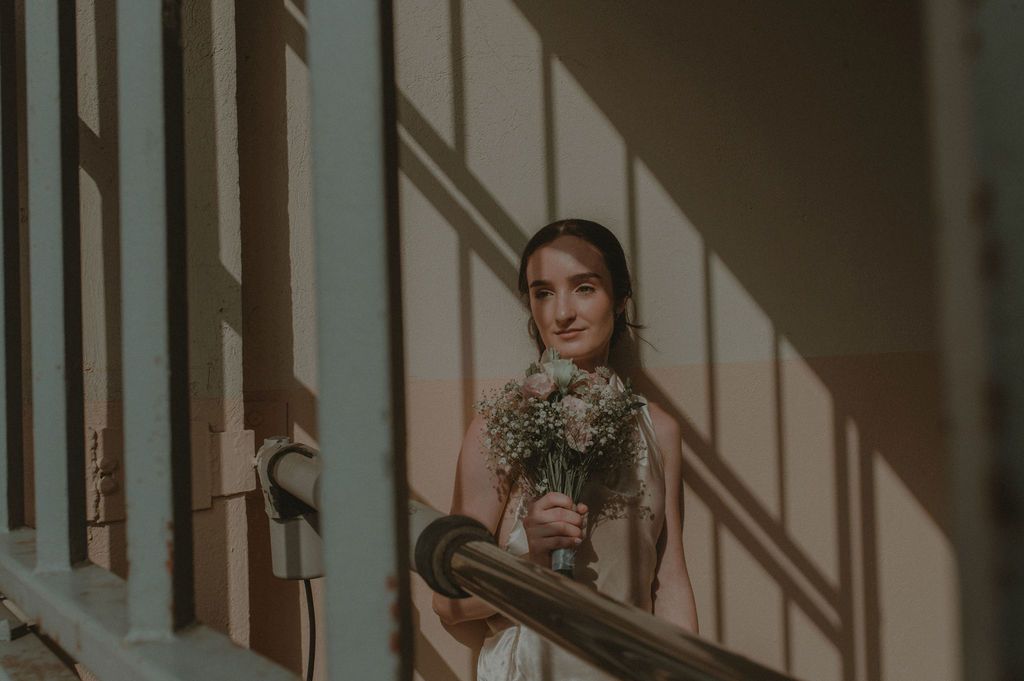 Bride in white dress holding small white bouquet stands on staircase with sunlight casting shadows on wall.