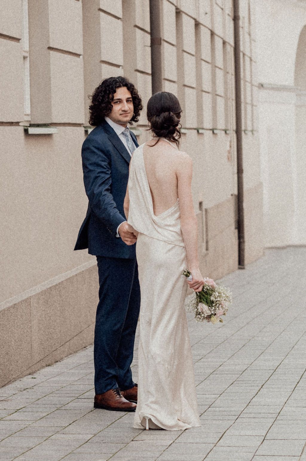 A couple in wedding attire holds hands on a cobblestone street, bride in backless gown holding bouquet, groom in navy suit.