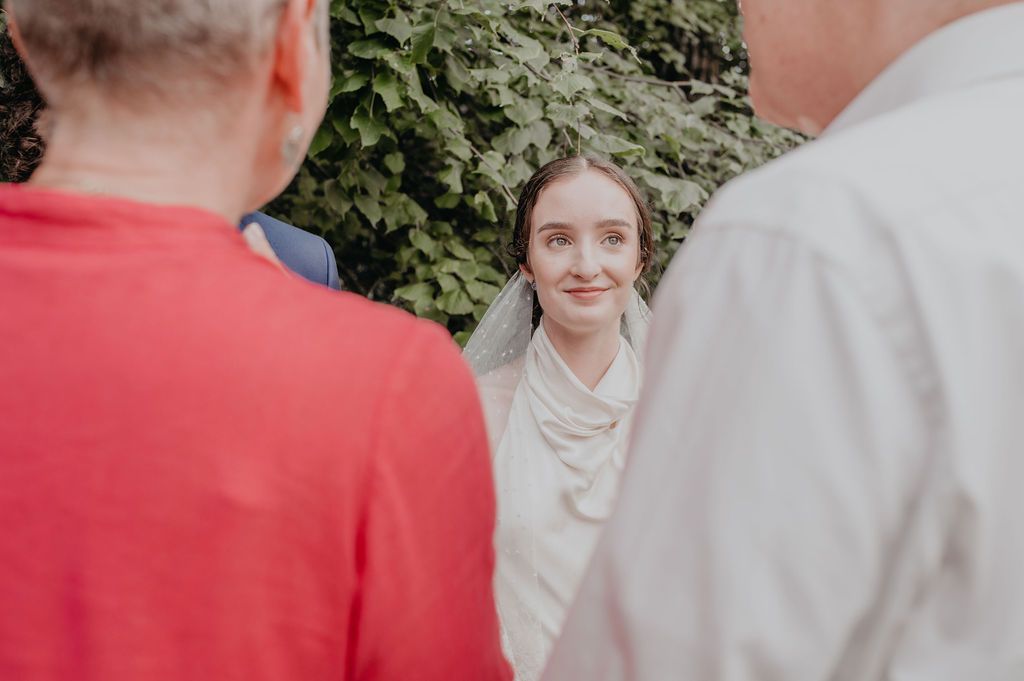 Beautiful bride in white gown and veil smiling warmly at wedding guests during outdoor ceremony.