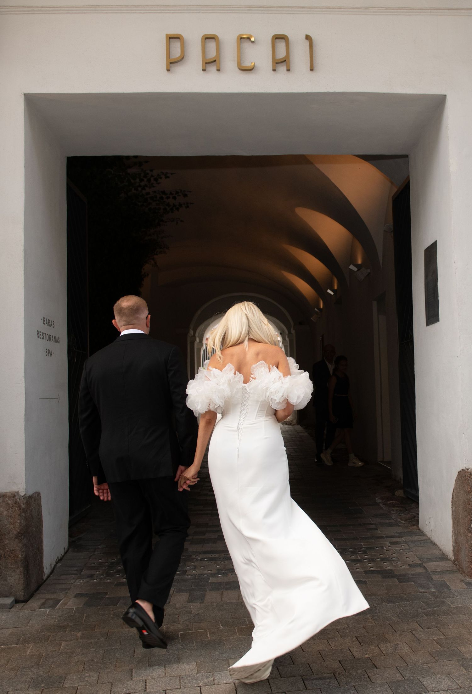 Bride in a white off-shoulder gown with ruffled sleeves holds hands with a groom in a black suit walking through a Pacai archway.