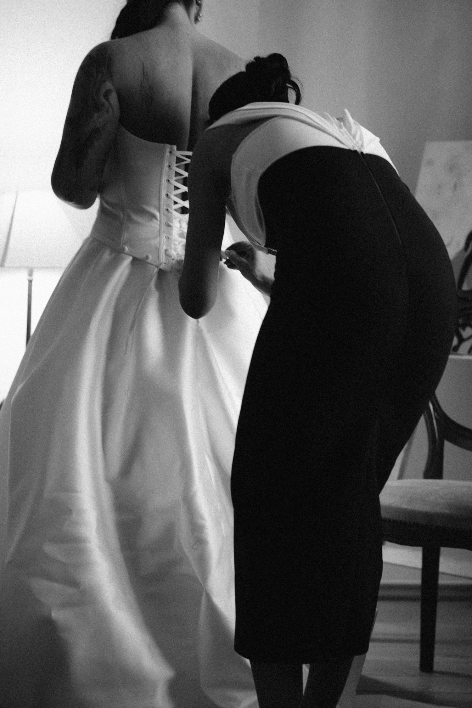 Black and white photo of a bridesmaid in a black dress lacing the corset back of a bride's white wedding gown before the ceremony.