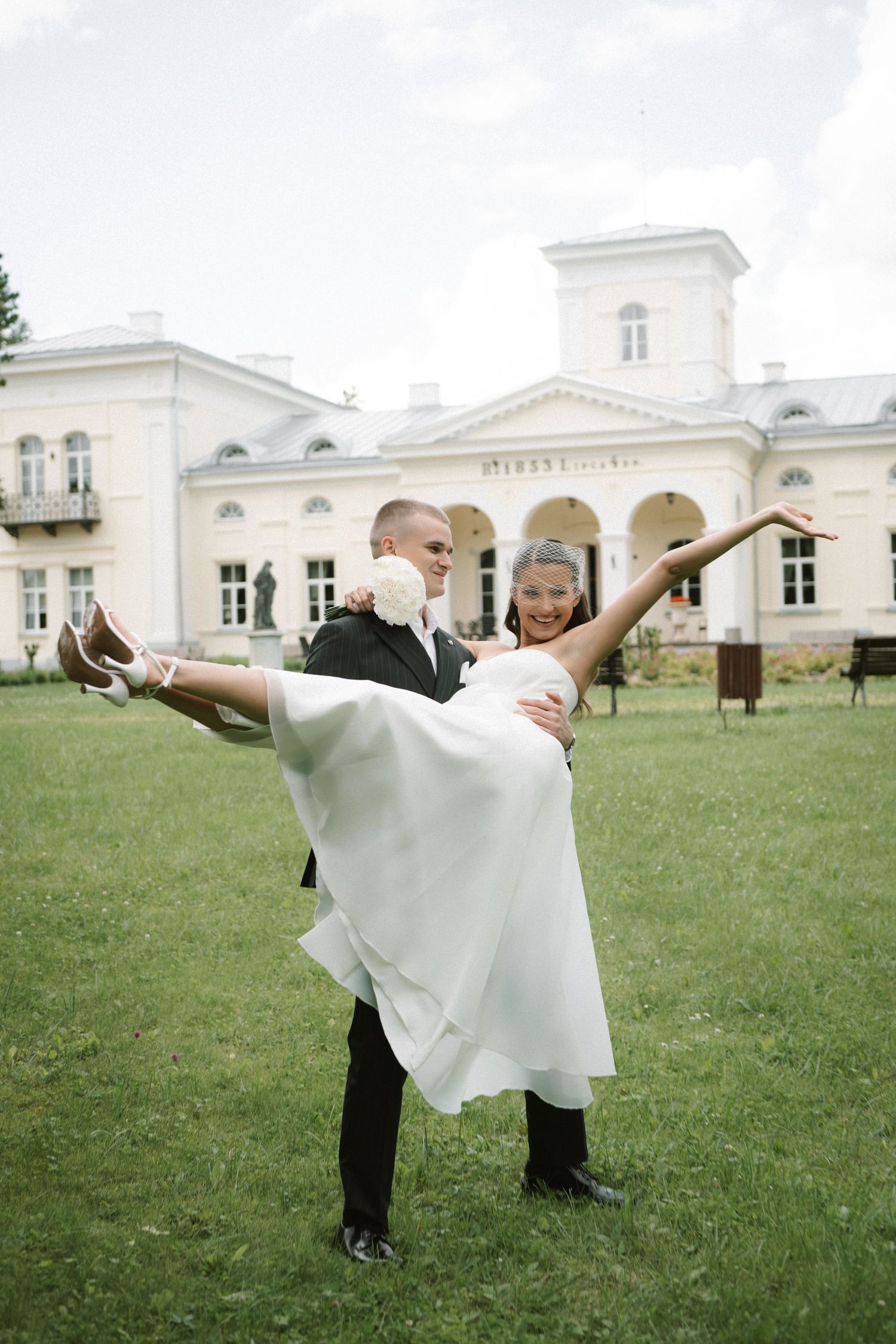 Groom in a dark suit lifts his smiling bride in a white dress into the air on a green lawn in front of a cream-colored manor house.