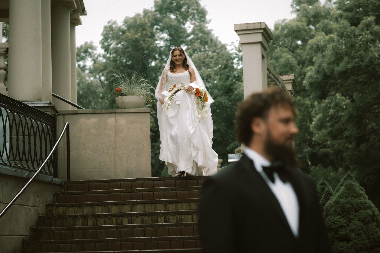 Bride in white gown descends stone steps while groom in tuxedo stands in foreground at outdoor wedding.