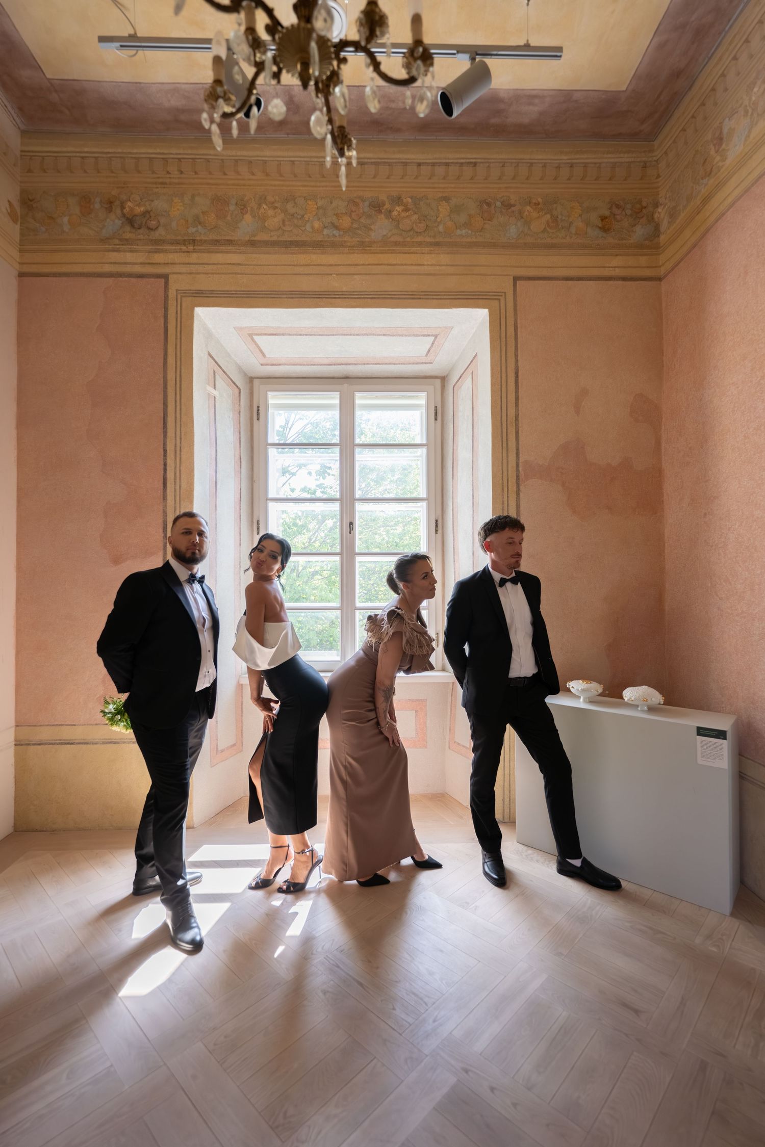 Four elegantly dressed guests in formal attire stand in a sunlit vintage room with ornate walls during a wedding event.