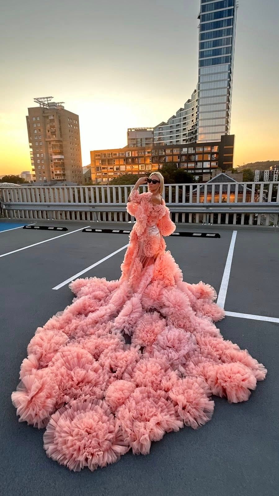 Woman in voluminous pink ruffled ball gown with long train poses on rooftop at sunset with city skyline.