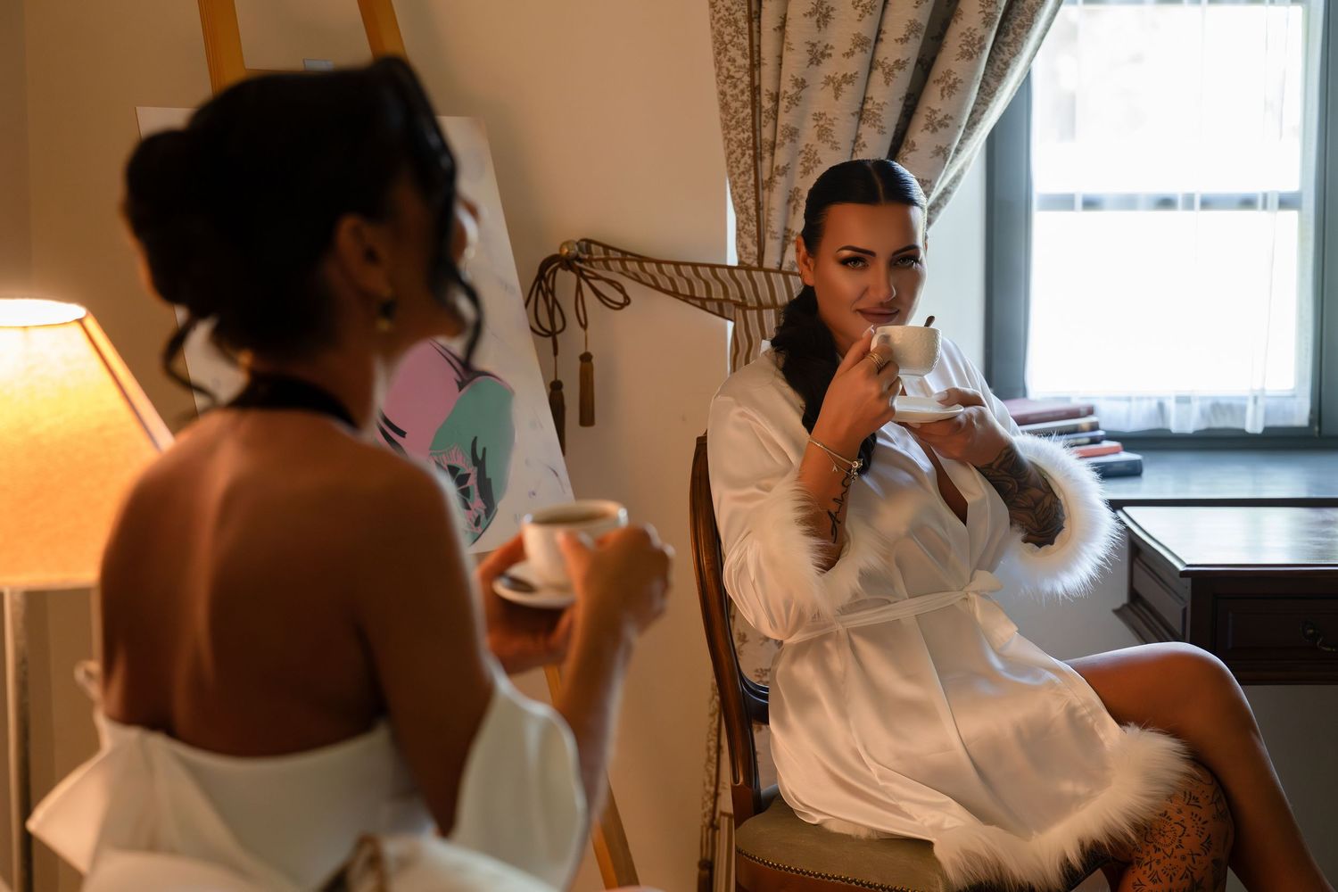 Two women in white satin robes with fur trim sip tea from porcelain cups in an elegant, softly lit room with draped curtains and a lamp.
