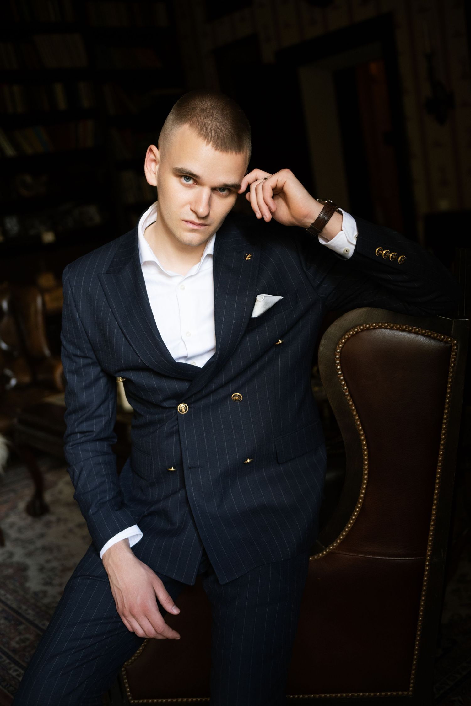 Young man in a navy pinstripe double-breasted suit and white pocket square sits in a brown leather armchair in a classic library.