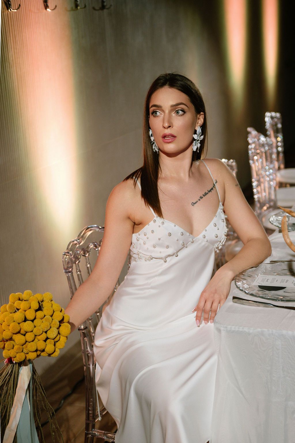Woman in a white satin slip dress with pearl details and crystal earrings sits at an elegant table beside a yellow billy ball bouquet.