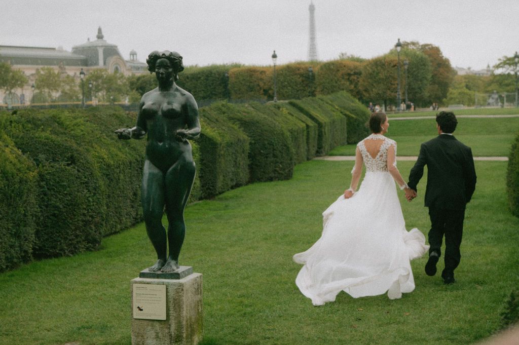 Bride and groom walk hand-in-hand through Tuileries Garden in Paris, with bronze statue and Eiffel Tower in background.