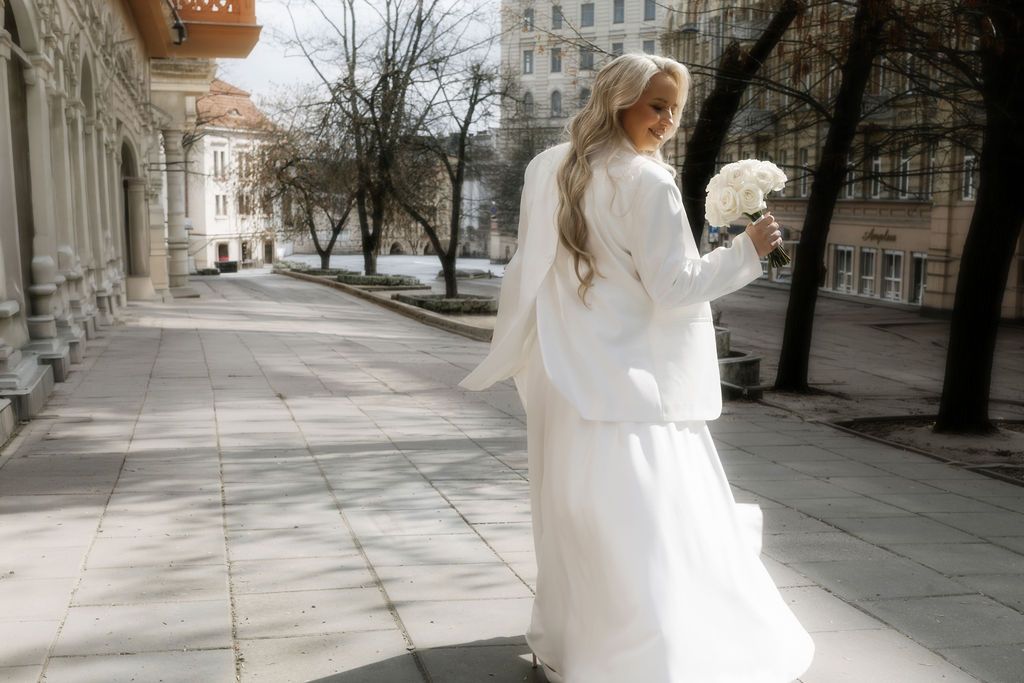 Elegant bride in white dress and jacket holding bouquet, walking along urban street with classic architecture.