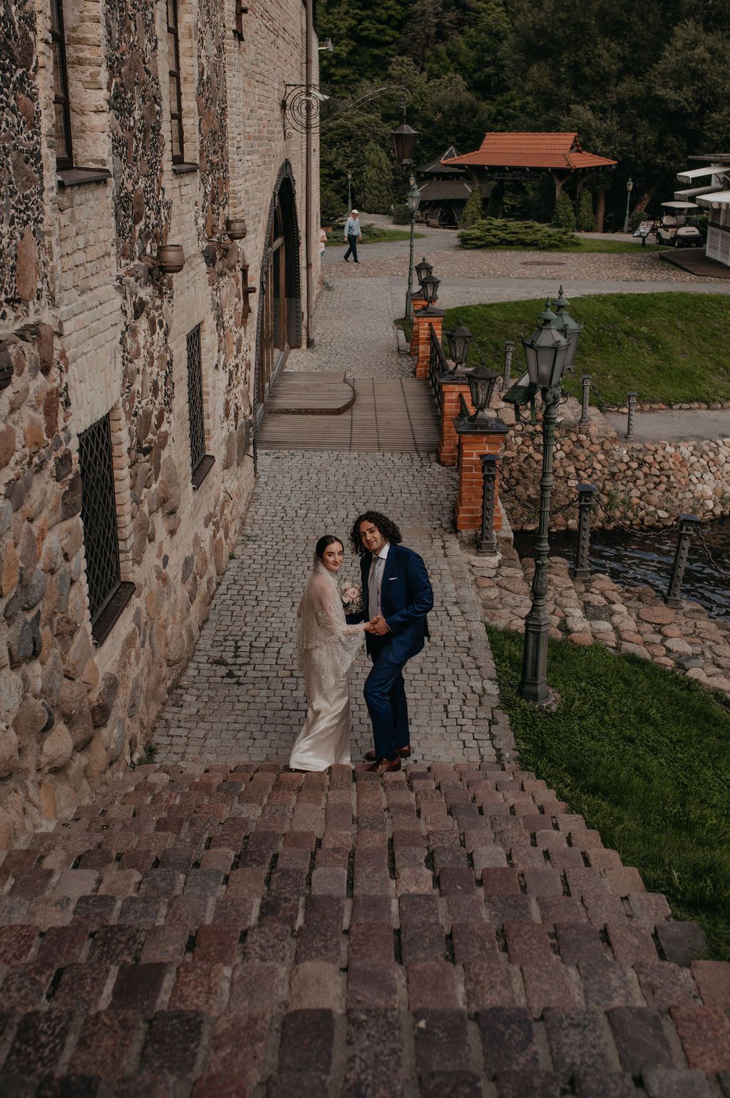 Bride and groom holding hands on cobblestone steps beside a rustic stone building at their wedding venue.