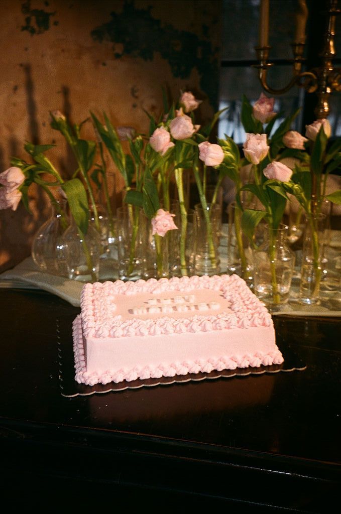 Pink square celebration cake with decorative frosting on dark table, surrounded by white roses in glass vases.