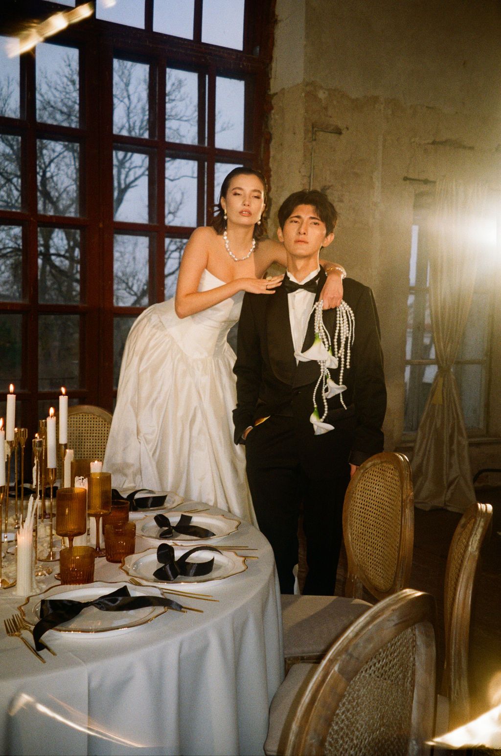 Elegant bride in white gown and groom in black tuxedo pose by candlelit reception table in rustic venue.