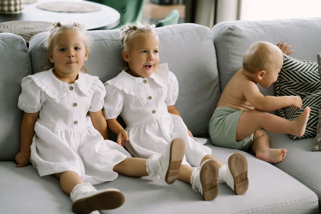 Three young children in white outfits sit on a gray sofa, two toddler girls in dresses and a baby in green shorts.